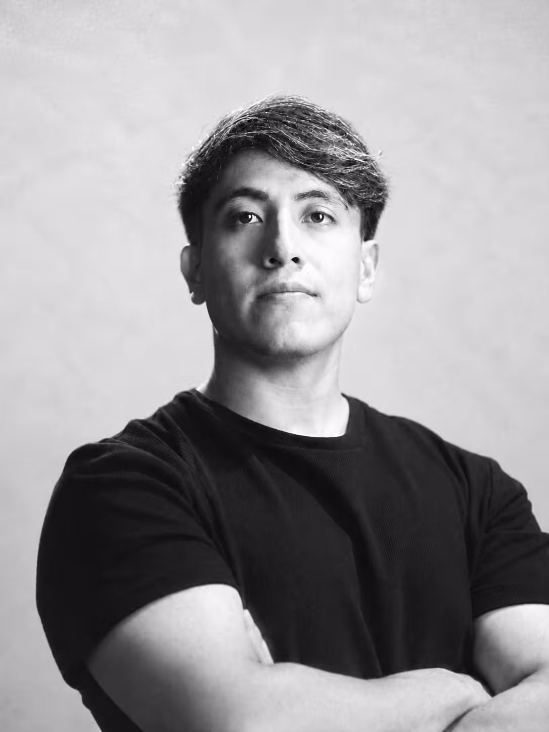Retrato profesional en blanco y negro de un hombre joven con los brazos cruzados, vistiendo playera negra, mirando ligeramente hacia arriba