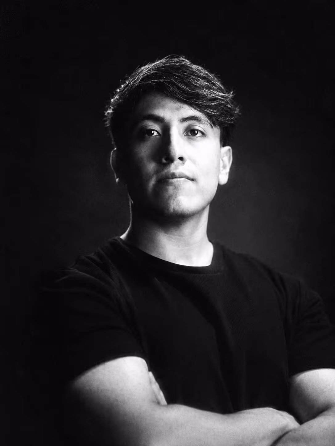 Retrato profesional en blanco y negro de un hombre joven con los brazos cruzados, vistiendo playera negra, mirando ligeramente hacia arriba